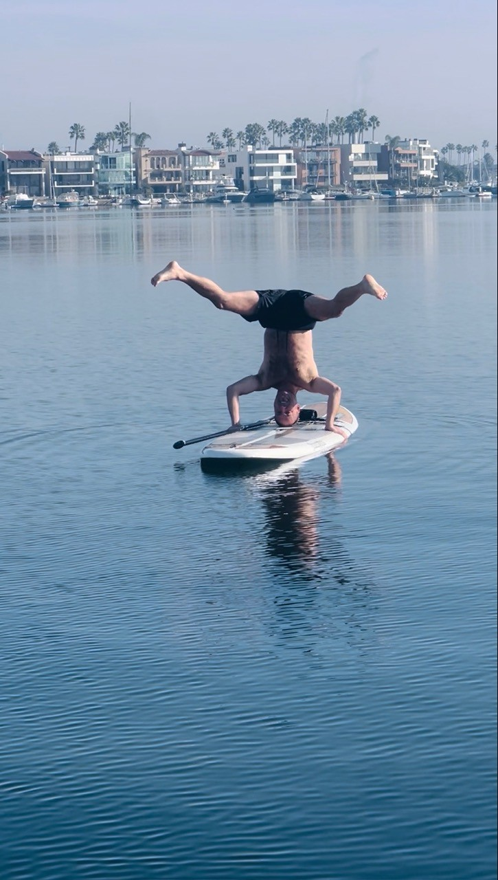 Rob Steven Williams holding a headstand on a paddleboard on calm bay water